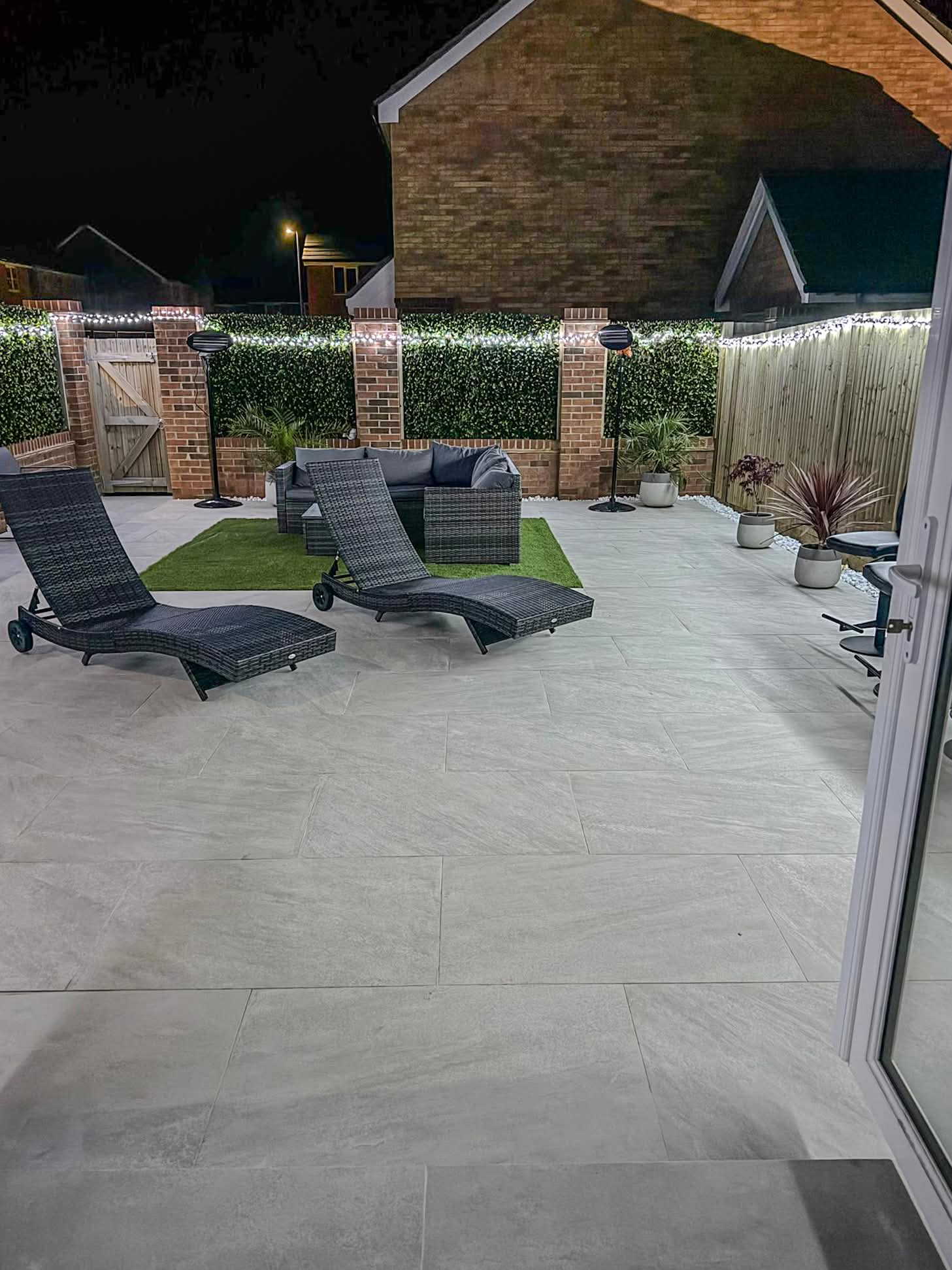 Finished grey porcelain patio at night with loungers, turf inset and festoon lighting